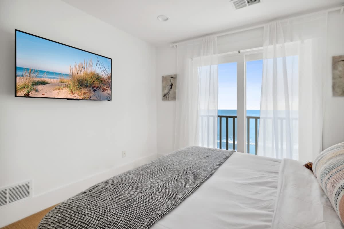 View of bedroom-1 showing a mounted TV on the wall opposite the bed, with a private balcony providing clear lake views. The room features a gray throw blanket and white sheer curtains, maintaining a minimalist aesthetic.