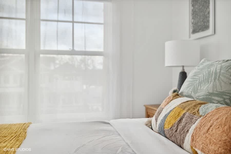 Close-up view of the bed in a bright bedroom, showing decorative pillows with botanical and geometric patterns, and a large window with sheer white curtains.