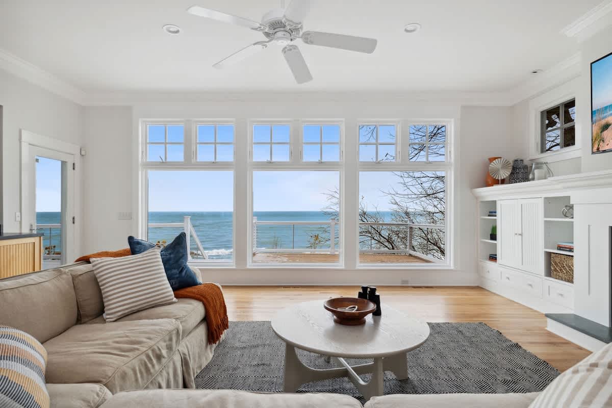 Bright living room with a large sectional sofa, a round coffee table, and expansive windows offering a clear view of the lake. A white fireplace with a mounted TV anchors one wall, and hardwood floors extend throughout.