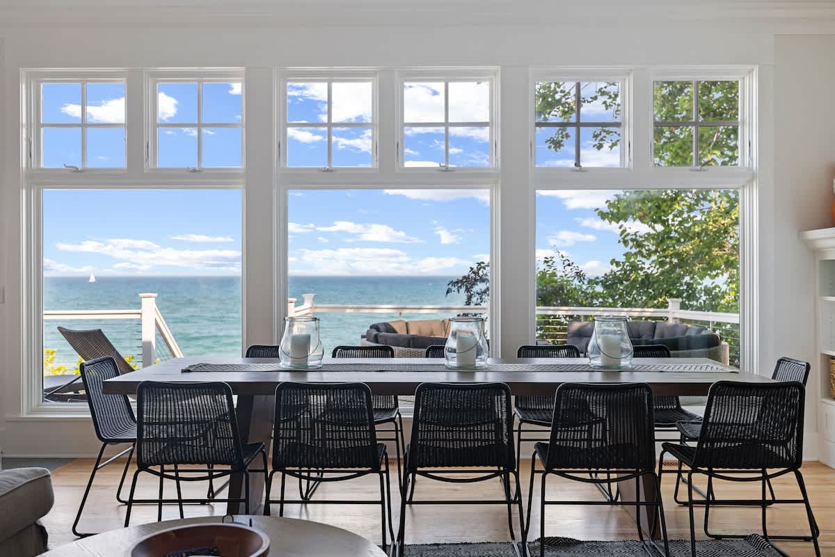 A spacious dining area features a long wooden table with black chairs, positioned in front of a wall of large windows that offer breathtaking, unobstructed views of the lake and sky.