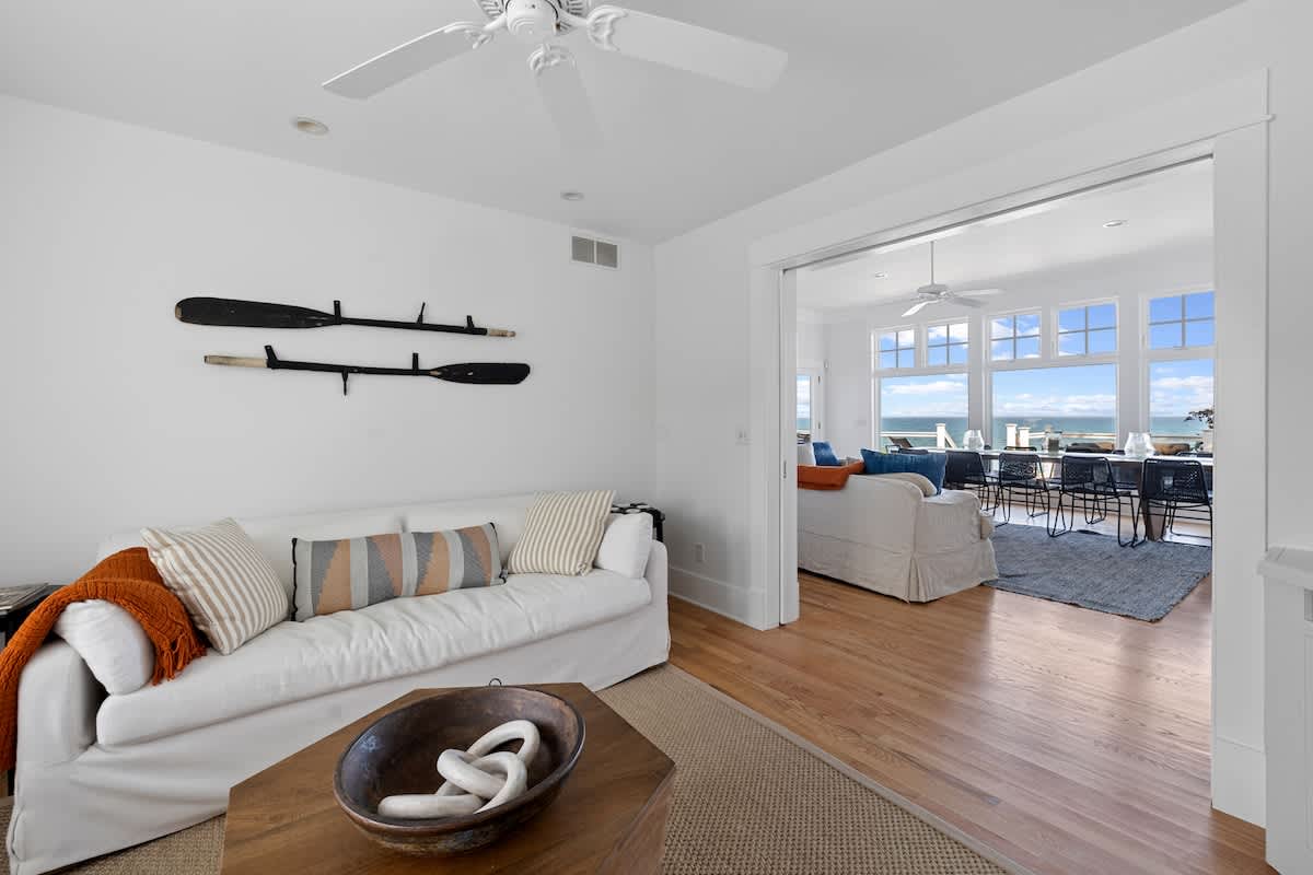 Bright living room featuring a white slipcover sofa with striped pillows, a rustic wooden coffee table, and a view through an open doorway to a dining area with expansive lake views. Decorative oars are mounted on the white wall.