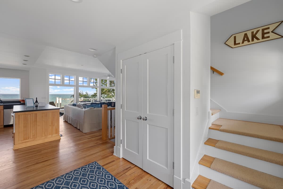 A bright foyer features light hardwood floors, a white staircase with carpeted treads, and a "LAKE" sign, opening into an open-concept living area with lake views.