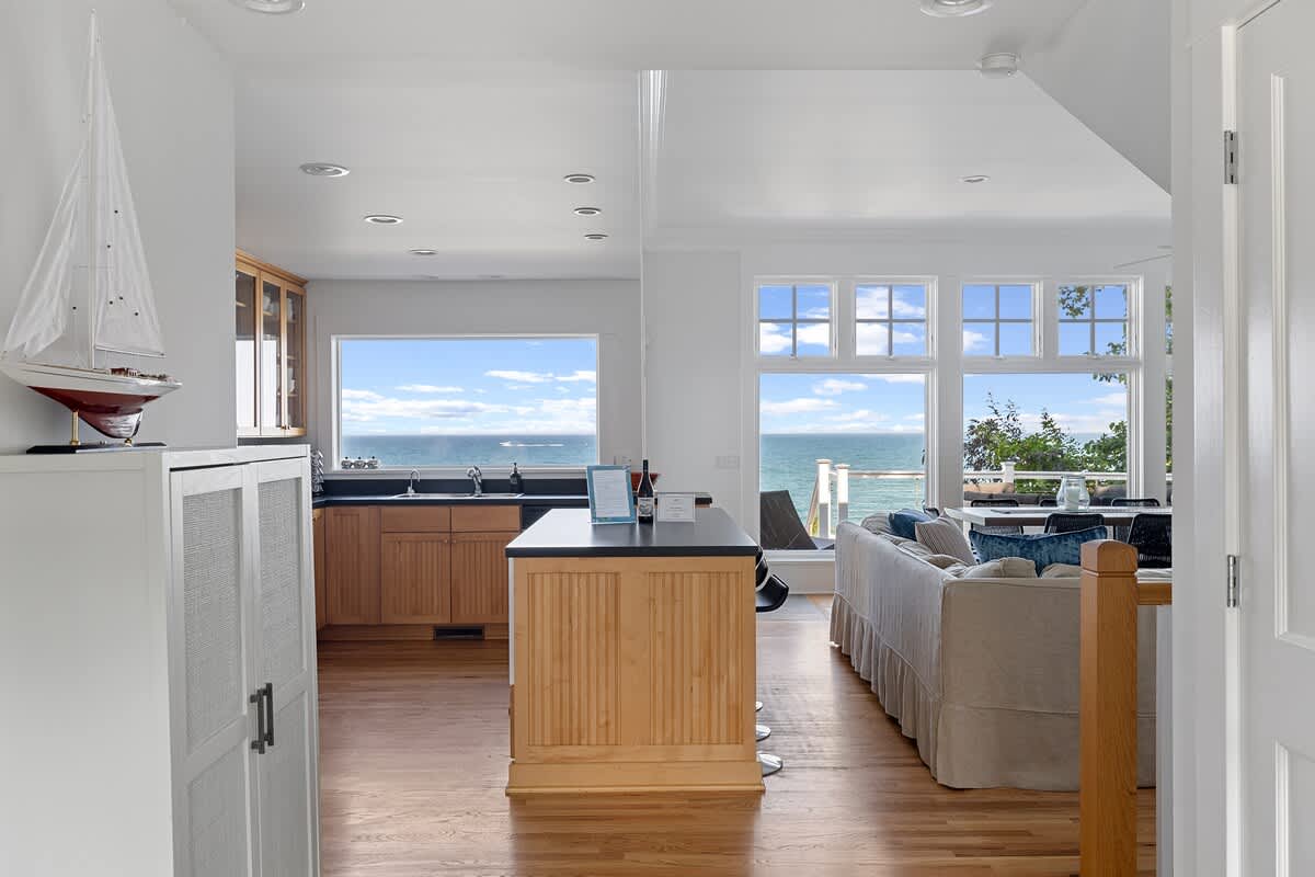 An open-concept space showcases a kitchen with light wood cabinetry and a dark island, flowing into a living area with a beige sofa, all overlooking expansive lake views through large windows. A decorative sailboat is visible on a cabinet.