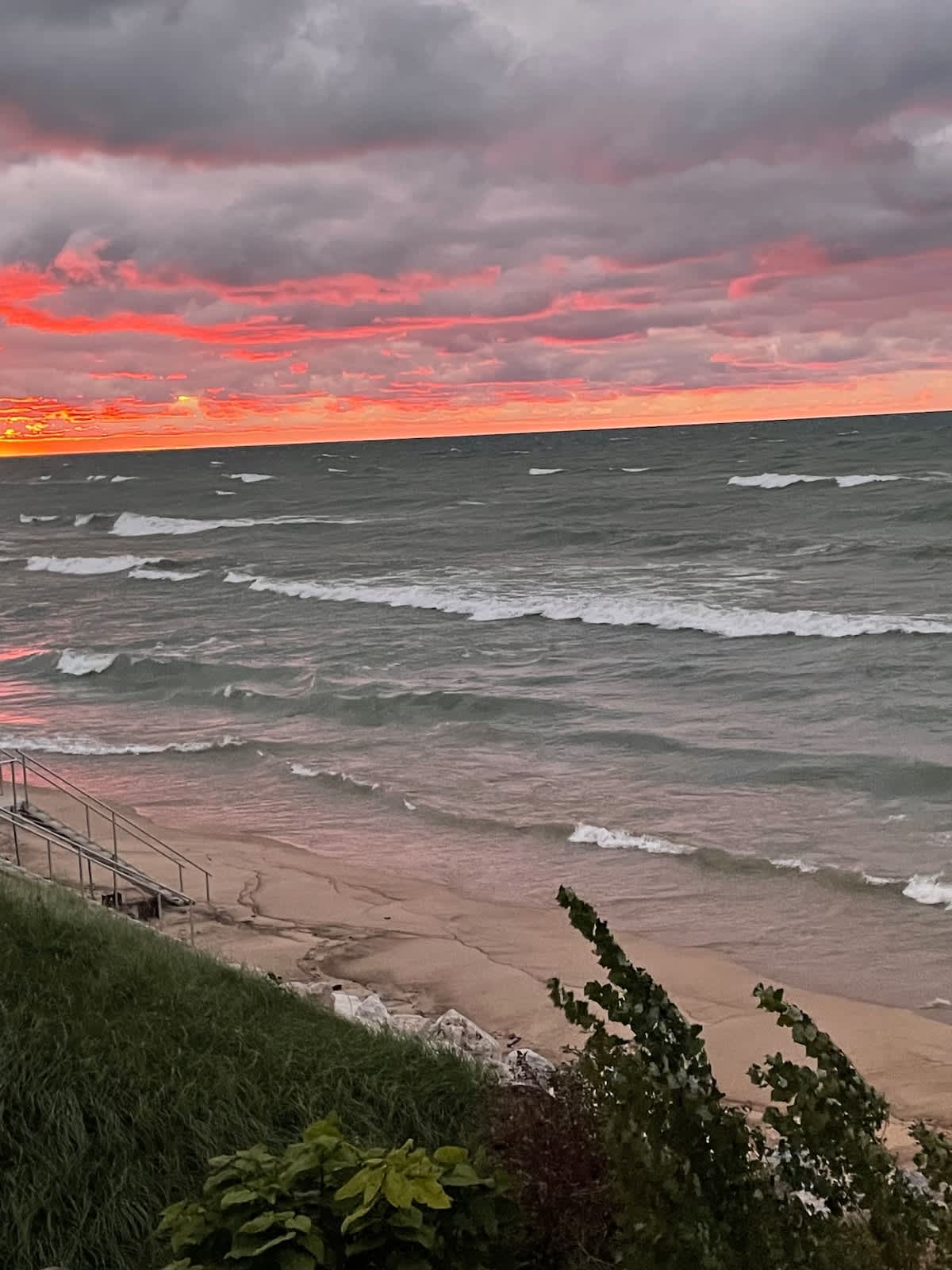 A dramatic sunset paints the sky with fiery orange and red hues over a vast, wavy lake, with a sandy beach and grassy dune in the foreground. Steps leading down to the beach are visible on the left.