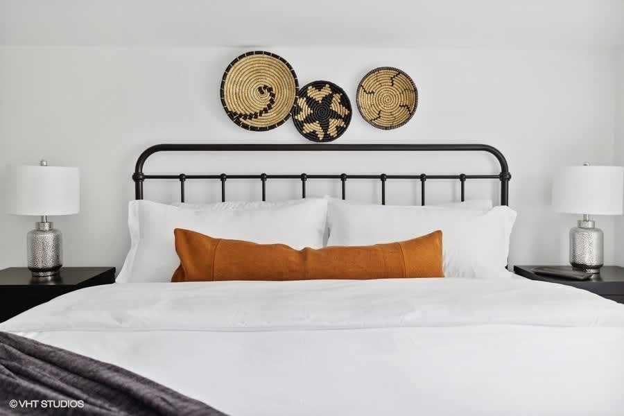 Bedroom with a black metal bed frame, white bedding, black nightstands with silver lamps, and three woven decorative baskets mounted on the white wall above the headboard.