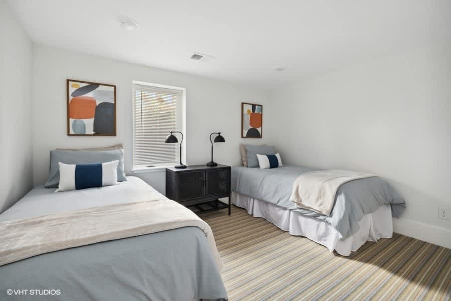 Bedroom with two twin beds, each with light blue bedding and decorative pillows, separated by a black cabinet with two black task lamps, and framed abstract art on the walls.