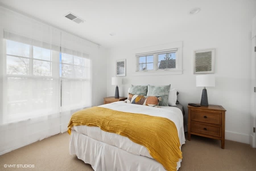 Bright bedroom featuring a white bed with a yellow throw blanket, two wooden nightstands with lamps, and large windows with sheer white curtains.