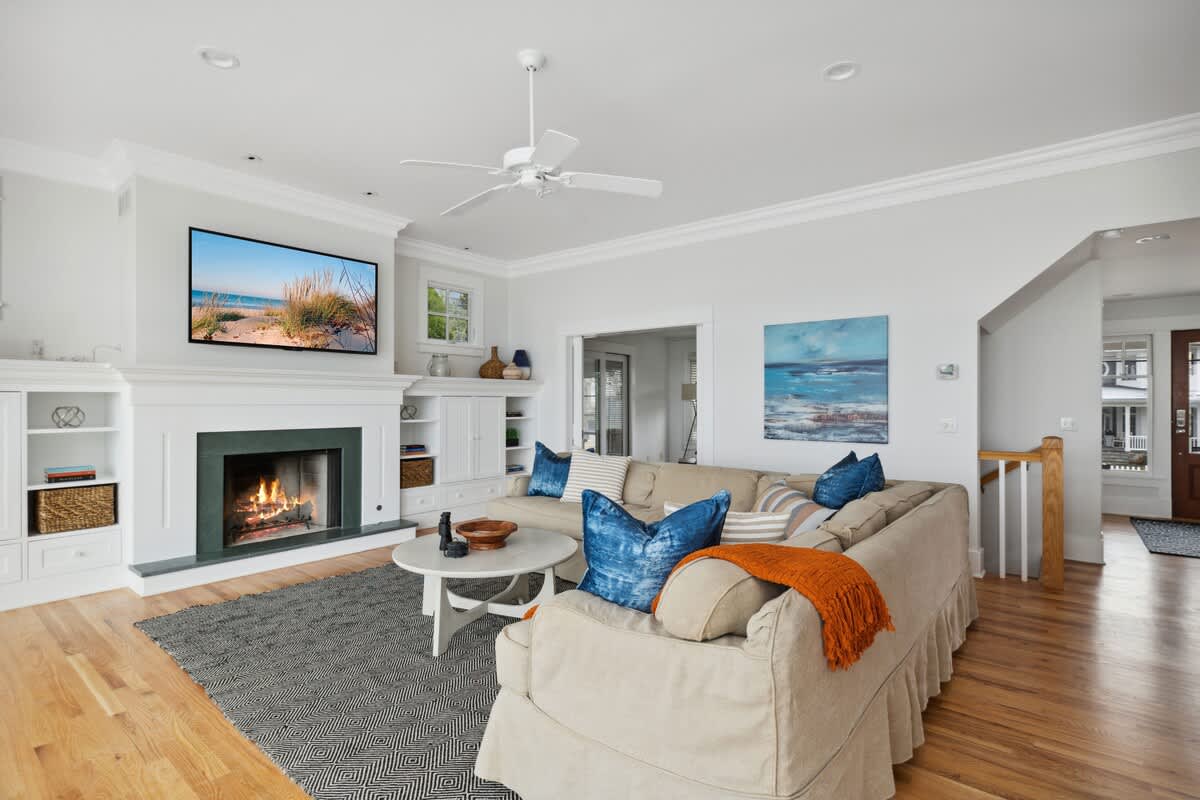 A spacious living room features a large beige sectional sofa with blue accent pillows, a white ceiling fan, and a fireplace with a mounted TV above, all set on a geometric patterned rug. Built-in shelves flank the fireplace.