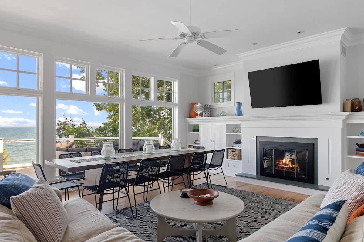 Bright open-concept living and dining area with large windows offering panoramic lake views. A long dining table with black chairs sits near the windows, while a cozy living area with a sectional sofa and fireplace is to the right.