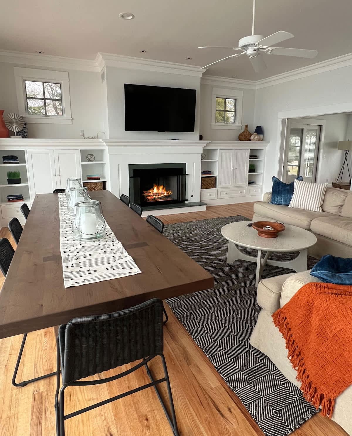 An open-concept space combines a dining area with a long wooden table and a living room featuring a fireplace, built-in shelving, and a large mounted TV, all on hardwood floors with a geometric rug.