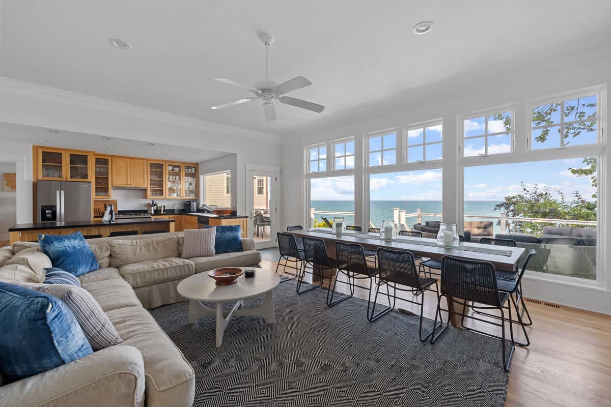 Expansive open-concept living space with a kitchen visible in the background, a dining area with a long table, and a living room with a large sectional sofa. Floor-to-ceiling windows offer stunning lake views.