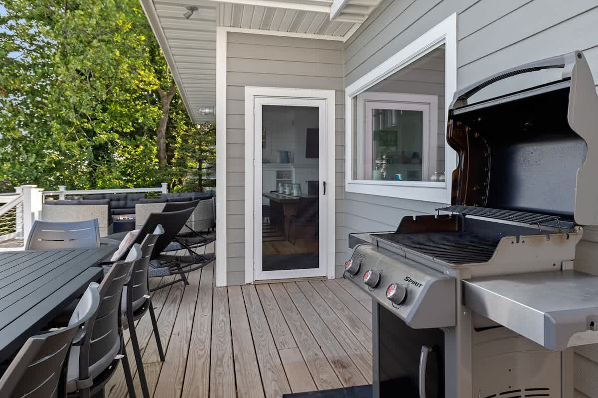A spacious wooden deck features a large stainless steel BBQ grill and a dark grey outdoor dining table with matching chairs. A glass-paned door leads into the house, and lush greenery is visible in the background.