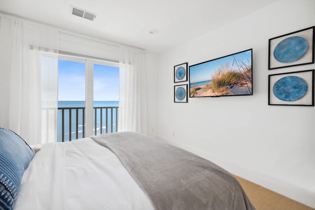 Another perspective of bedroom-4, highlighting the mounted TV and decorative blue wall art. The private balcony offers stunning views of the lake and horizon, framed by sheer white curtains.