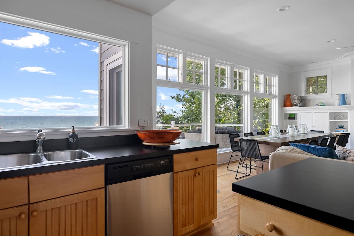 A bright kitchen area with light wood cabinets, black countertops, and stainless steel dishwasher, offers a panoramic lake view through a wide window, adjacent to a dining area with large windows.