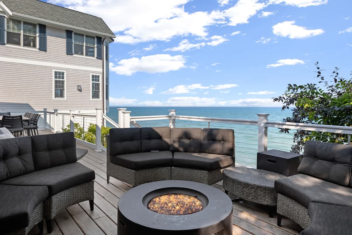 Spacious outdoor deck with a circular fire pit surrounded by comfortable dark gray wicker sectional seating, offering a direct view of the expansive lake under a bright blue sky.