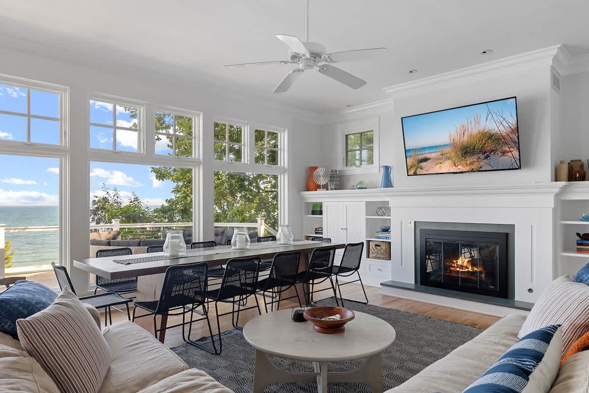 Open-concept living and dining area featuring a long dining table with black chairs by large windows overlooking the lake. A comfortable sectional sofa faces a fireplace with a mounted TV, all under a ceiling fan.