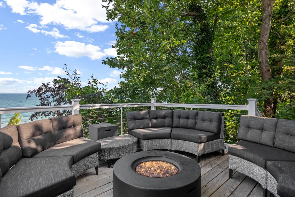 A curved outdoor sectional sofa in dark grey surrounds a modern circular fire pit on a wooden deck, offering a cozy gathering space. Beyond the railing, the vast blue lake and lush trees create a serene backdrop.
