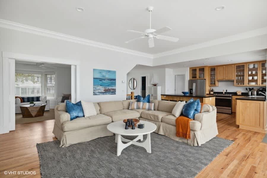 A bright living room features a large beige sectional sofa with blue pillows, a white coffee table, and a geometric rug, with a view into an adjacent sitting area and the kitchen beyond.