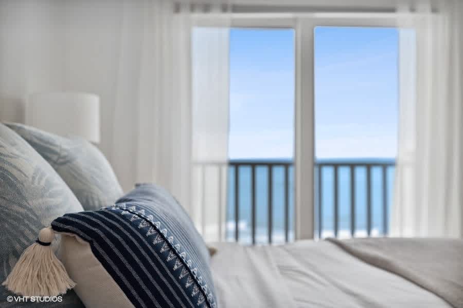 Close-up detail of decorative pillows on a bed, with a blurred background showing a private balcony and the expansive blue lake beyond. The pillows feature blue and white patterns, adding a coastal touch.