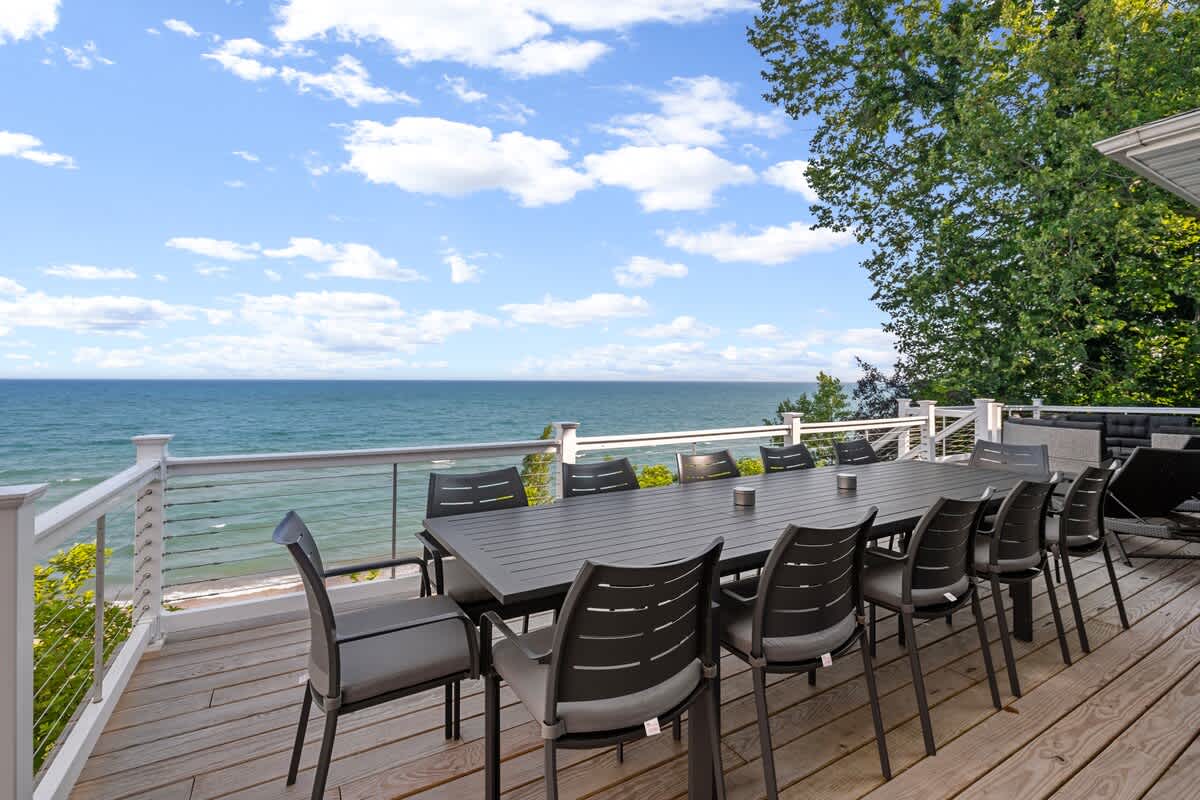 A spacious outdoor deck features a large dark dining table with multiple chairs, set against a stunning backdrop of a vast lake under a blue sky with scattered clouds.