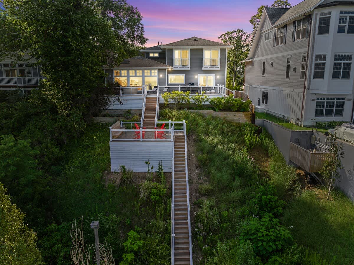 An aerial view showcases the multi-level house with extensive wooden decks and long stairs descending a grassy slope towards the lake. The property is surrounded by lush green trees under a vibrant sunset sky.