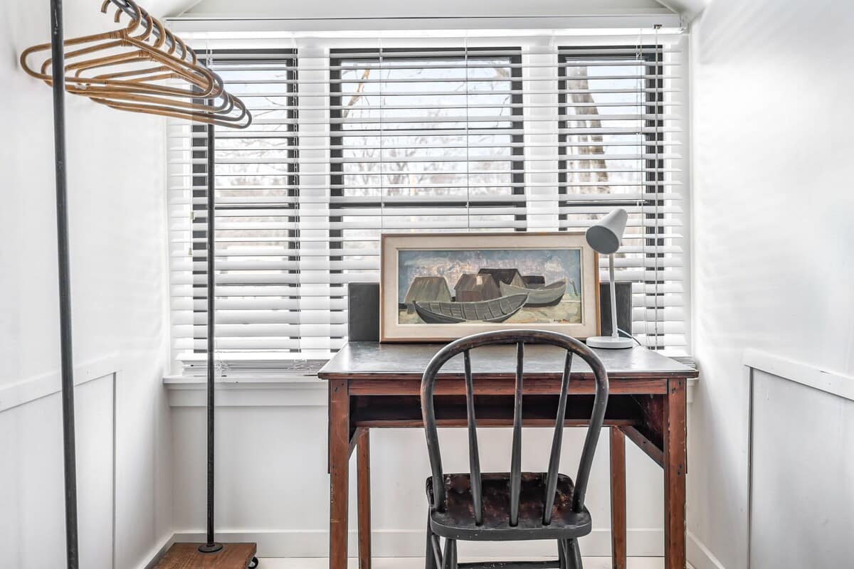 Close-up of a wooden desk with a framed painting of boats and a modern lamp, positioned by a window with white blinds. A dark wooden chair is tucked under the desk, and a metal clothes rack is visible.