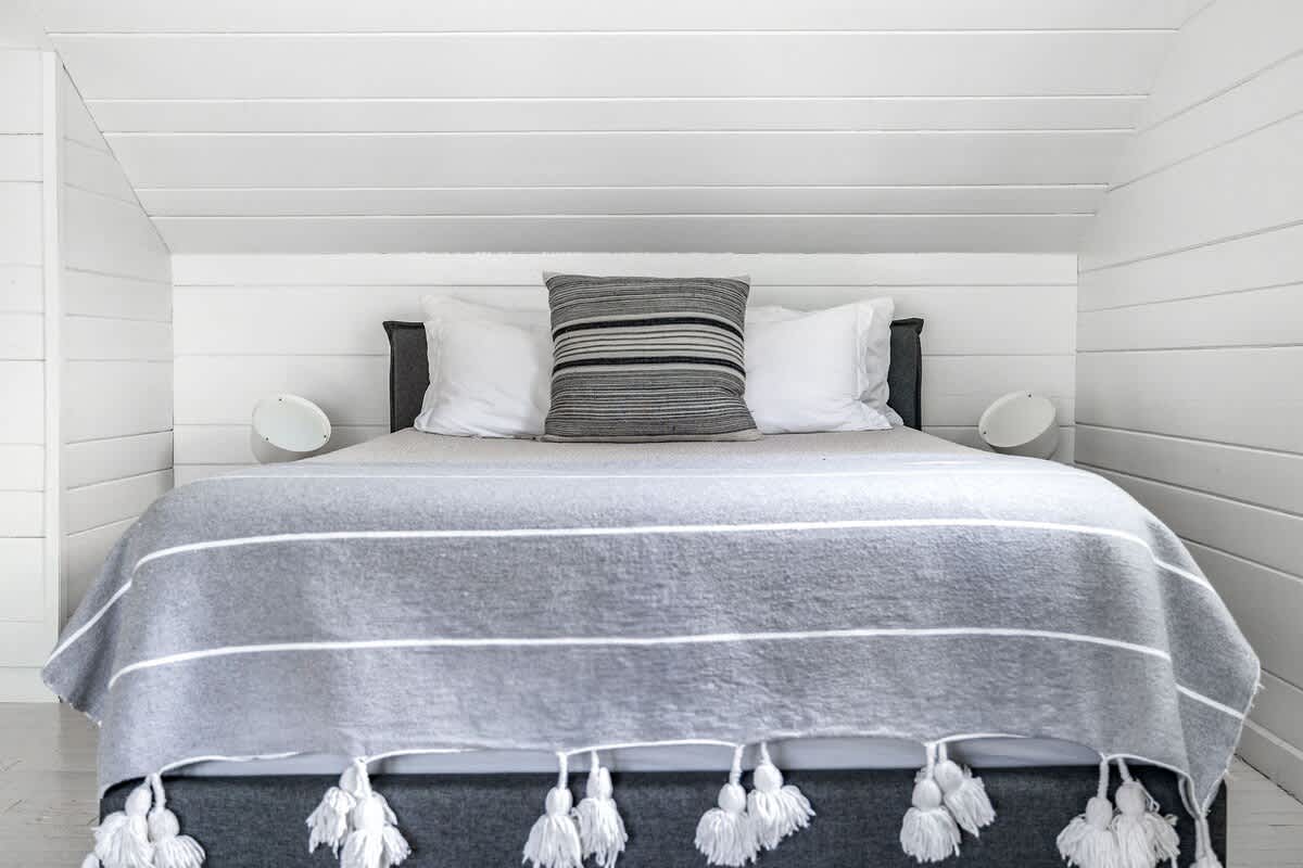 A close-up view of a bed in a bedroom with white shiplap walls, featuring a gray upholstered bed frame, white bedding, a gray striped throw with decorative tassels, and two white wall sconces.
