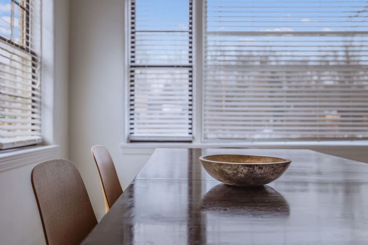Close-up of the dark wood dining table with a decorative bowl, showing two modern dining chairs and large windows with horizontal blinds in the background.