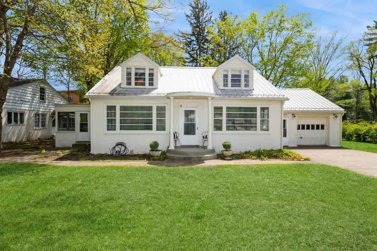 Charming white cottage with a light-colored metal roof and two dormer windows, a small covered front porch, and a well-manicured green lawn leading to a concrete driveway.