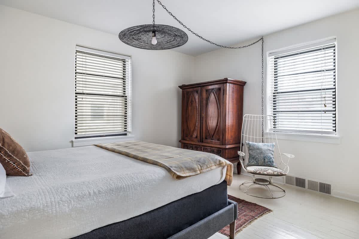 A bright bedroom with white wooden floors and walls, featuring a dark gray upholstered bed, two windows with white blinds, a dark antique wooden wardrobe, and a modern white hanging chair.
