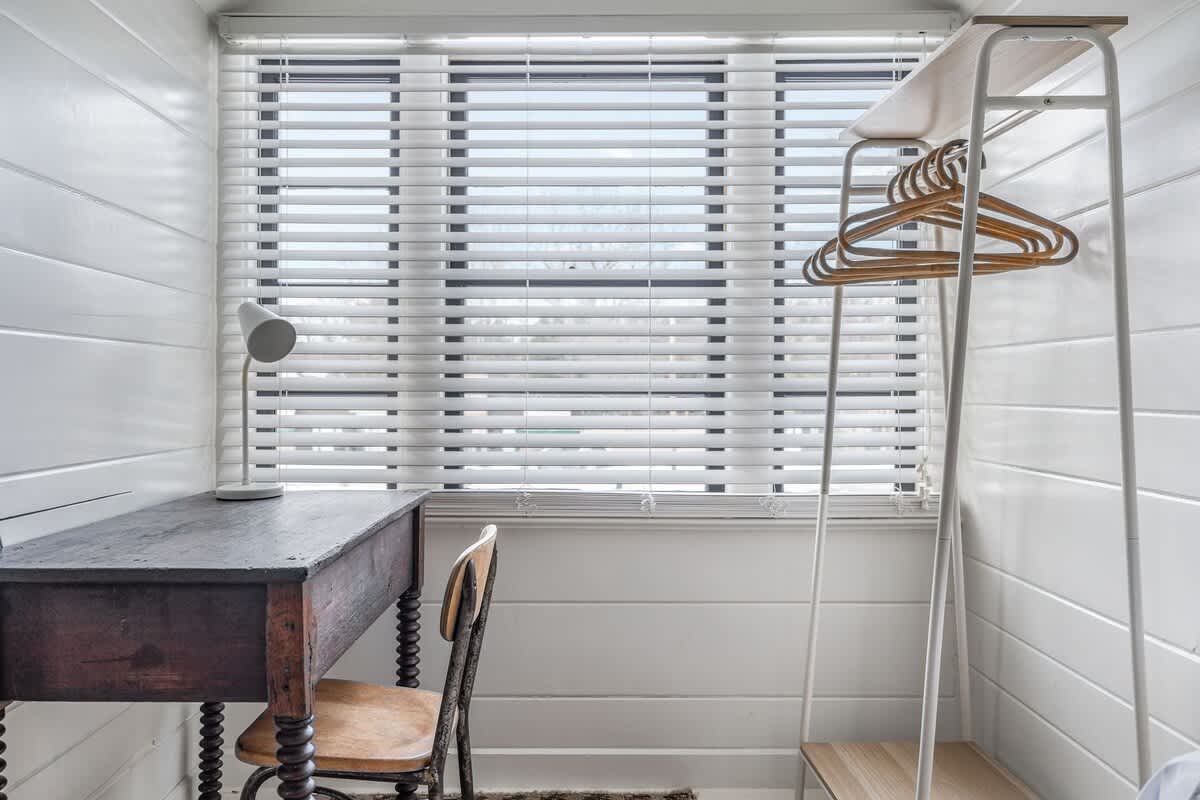 A dedicated workspace in an attic bedroom with white shiplap walls, featuring a dark wooden desk with a vintage chair, a white table lamp, and a minimalist white clothing rack next to a window with blinds.