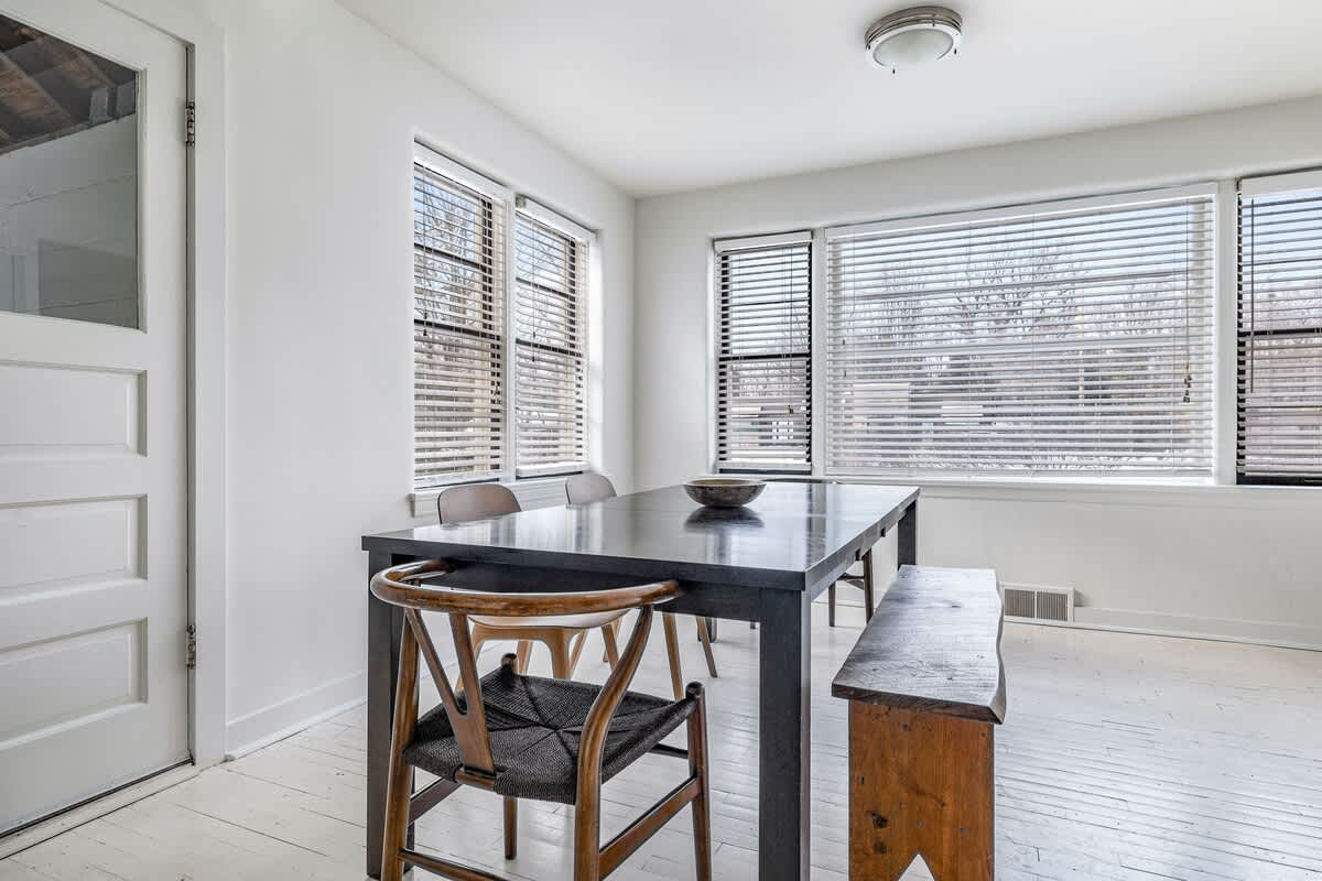 Dining area featuring a large dark wood table with mixed chairs and a wooden bench, surrounded by multiple windows with blinds, and light-colored hardwood floors.
