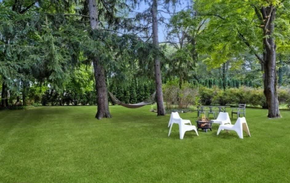 Lush green backyard featuring a circular fire pit area with four modern white chairs on a gravel base, surrounded by tall trees, with a hammock visible in the background.