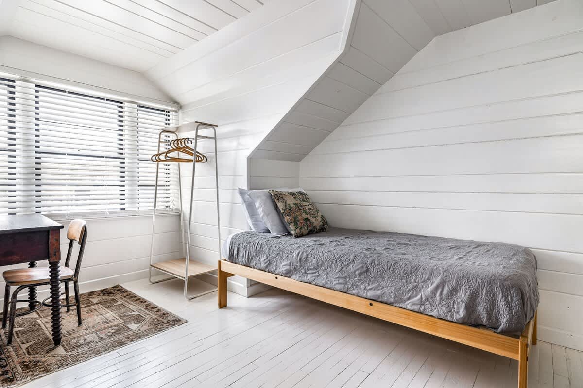 Attic bedroom with white shiplap walls and floorboards, featuring a single bed with a grey patterned quilt, a wooden desk and chair by a window with blinds, and a metal clothes rack.