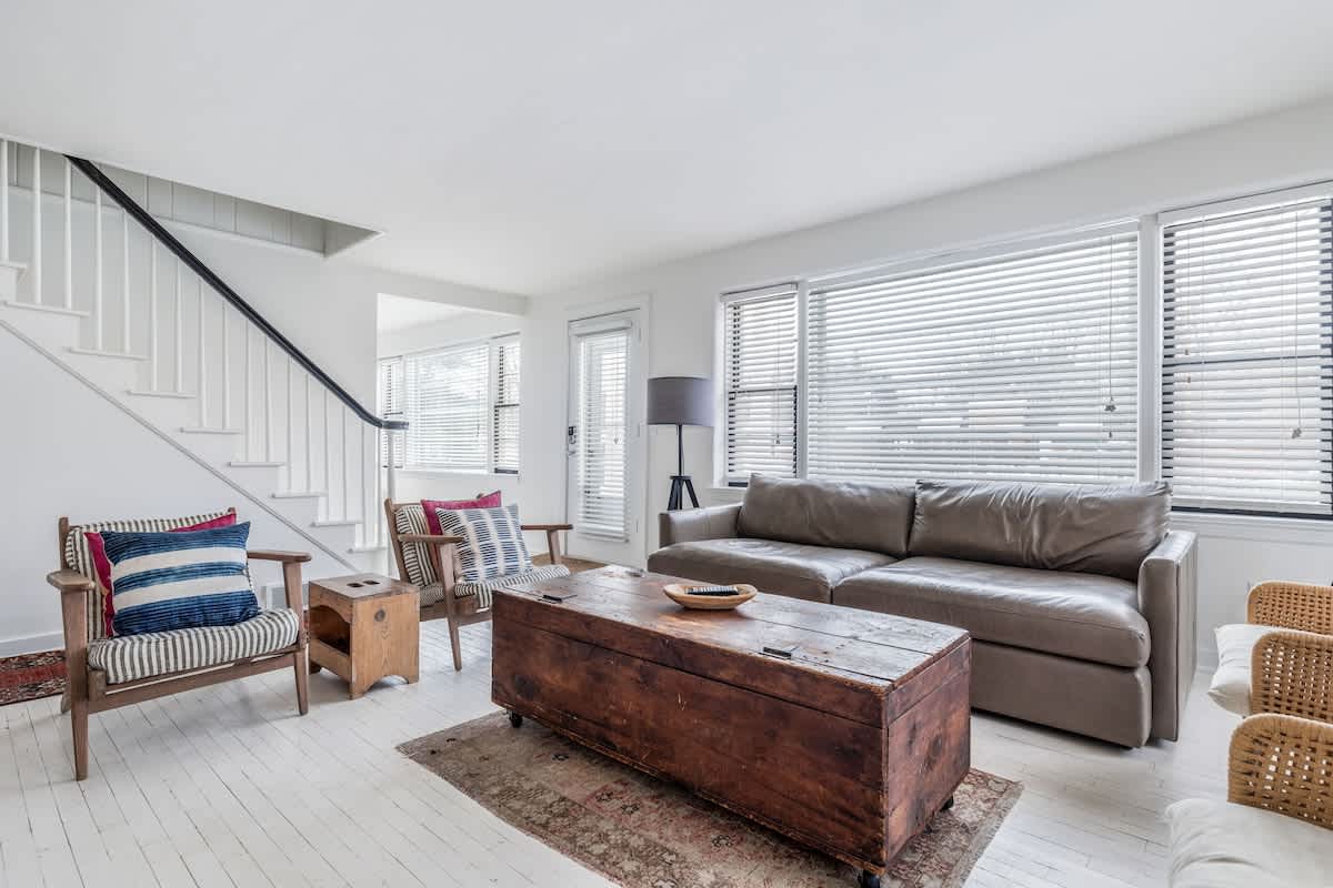 Spacious living room with white painted wood floors, a large brown leather sofa, and various armchairs. A rustic wooden trunk coffee table sits on an area rug, and a white staircase with a black handrail is visible in the background.