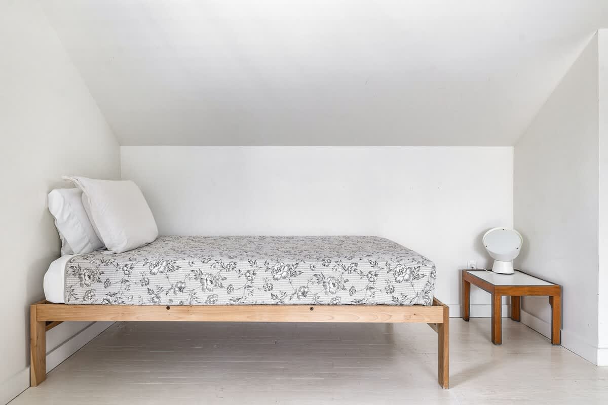 Minimalist attic bedroom with white walls and painted floorboards, featuring a single bed with a light floral patterned quilt and a small wooden side table with a modern circular lamp.