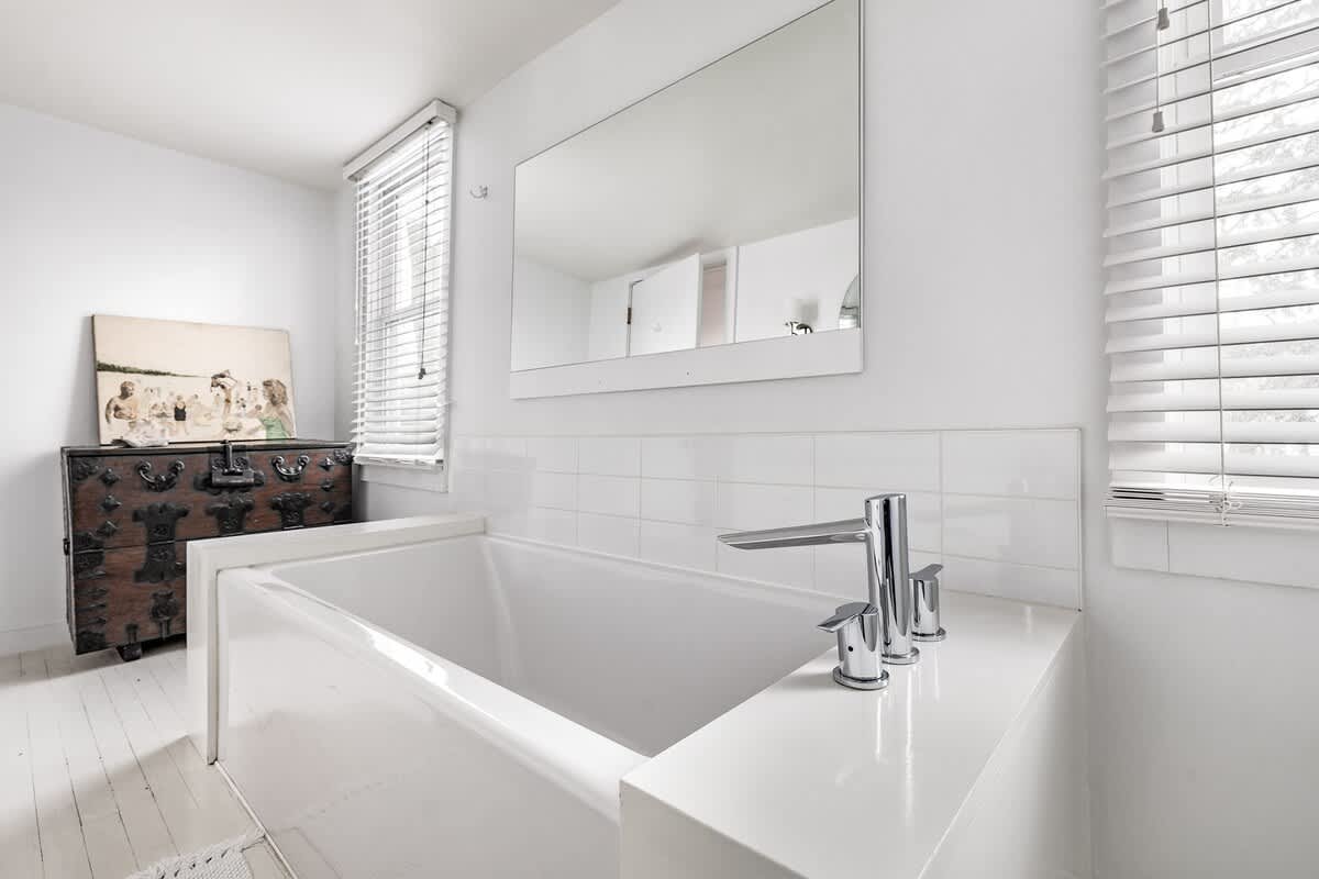 Bright white bathroom featuring a deep soaking tub with chrome fixtures, a large rectangular mirror above, and a decorative wooden chest with artwork nearby. A window with blinds is visible.