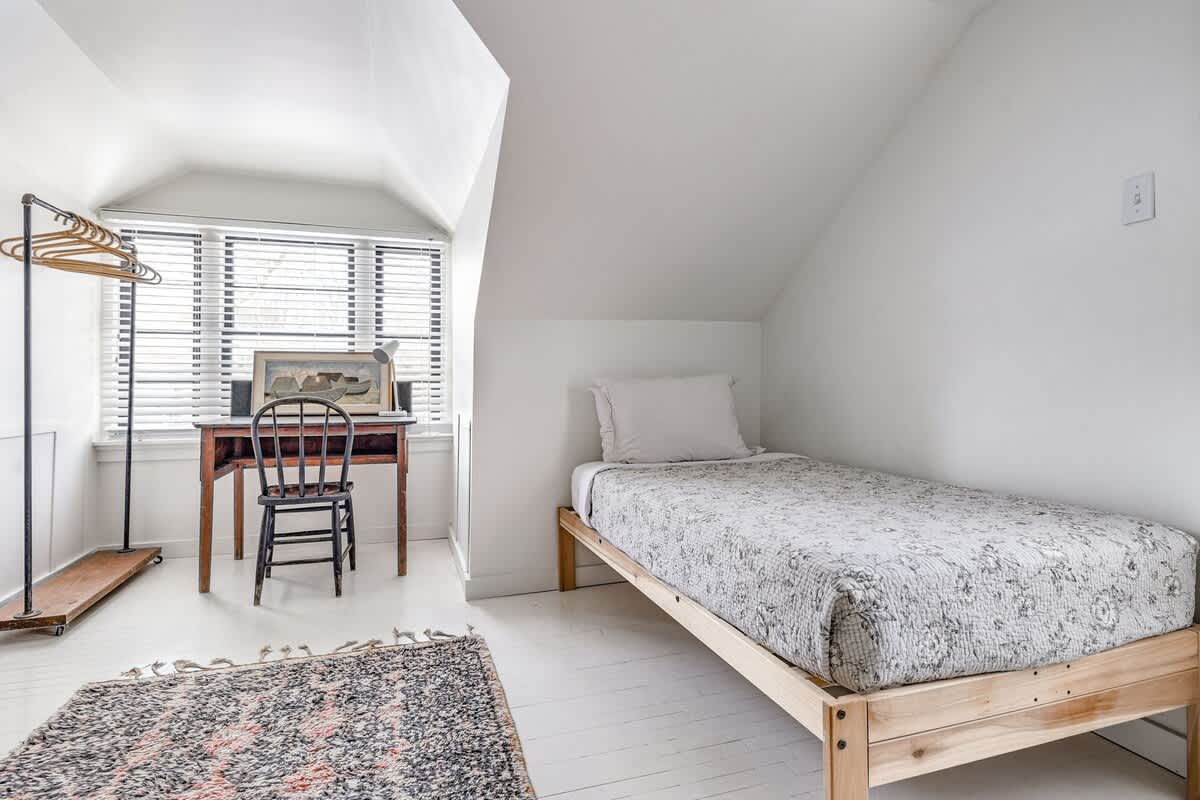 Bright attic bedroom nook with white walls and painted floorboards, featuring a single bed with a patterned quilt, a wooden desk and chair by a window with blinds, and a metal clothes rack.