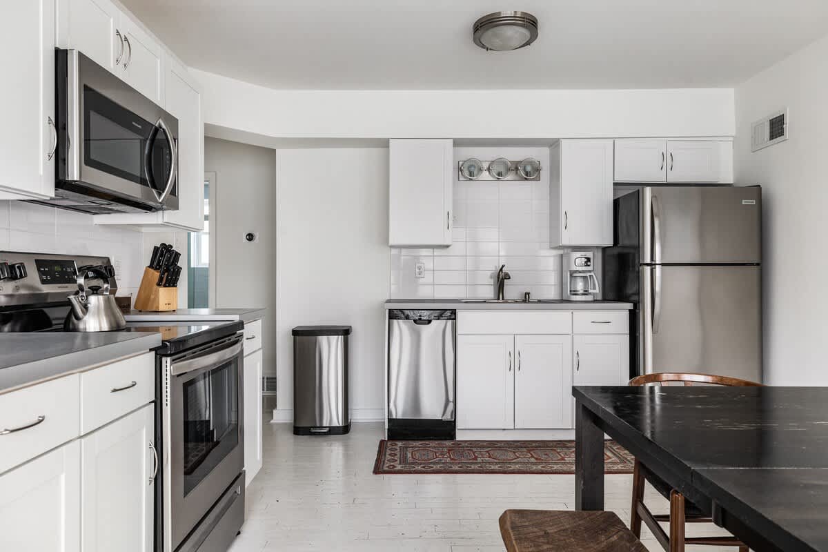 Bright kitchen featuring white shaker cabinets, stainless steel appliances including a microwave, range, dishwasher, and refrigerator, with light gray countertops and a dark wood dining table.