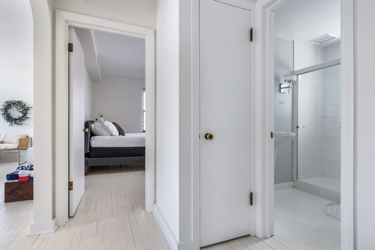 A bright, minimalist hallway with white wooden floors and walls, featuring open doorways leading to a bedroom on the left and a bathroom with a glass-enclosed shower on the right.