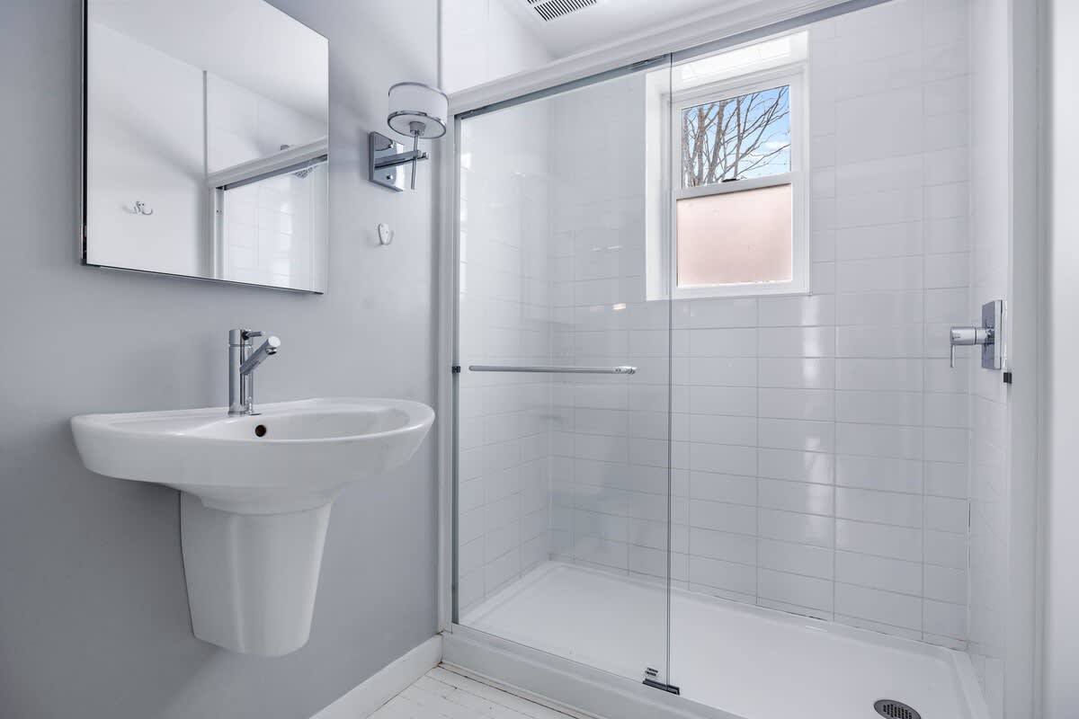 A modern bathroom with light gray walls, a white pedestal sink with a chrome faucet, and a glass-enclosed shower with white subway tiles and a window providing natural light.