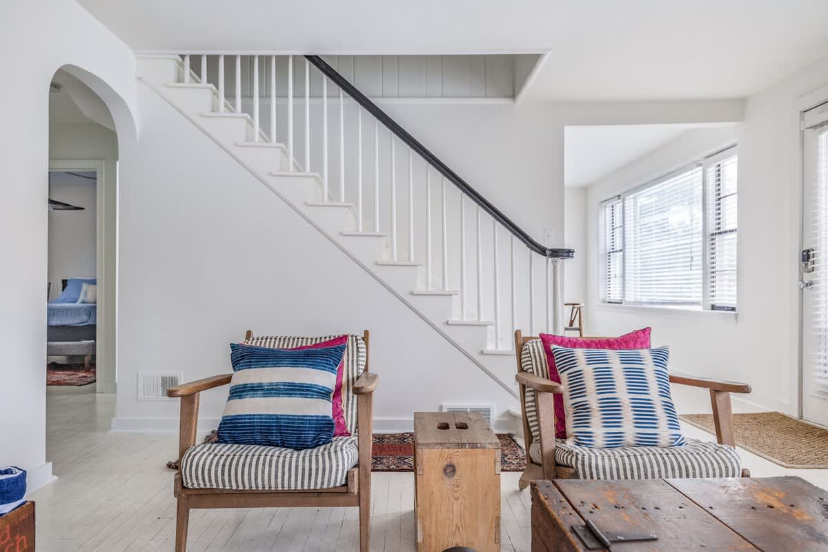 View of a white staircase with a black handrail, leading upstairs from a bright living area with white wood floors. Two armchairs with striped cushions are visible in the foreground, and an arched doorway leads to another room.
