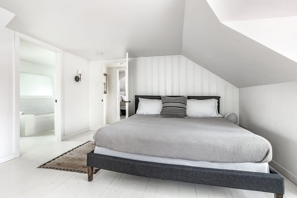 Cozy attic bedroom area with a queen-sized bed featuring a dark grey upholstered frame and a light grey textured blanket, with white walls and painted floorboards. A doorway leads to an adjacent bathroom.