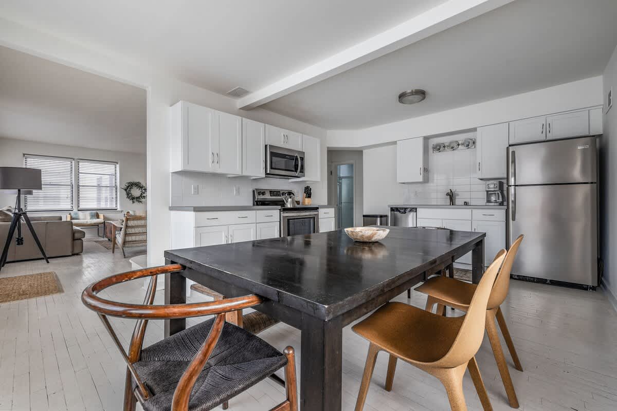 Open concept kitchen and dining area with white cabinets, stainless steel appliances, a large dark wood dining table, and light-colored hardwood floors extending into a visible living space.