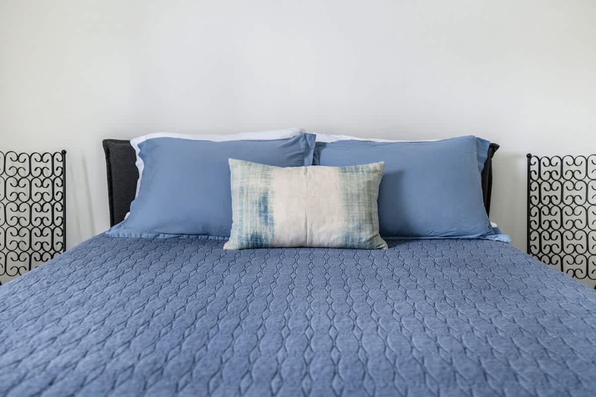 Close-up of the queen bed in bedroom-1, featuring blue pillowcases, a light blue and white decorative pillow, and a blue quilted comforter, with black metal headboard accents.