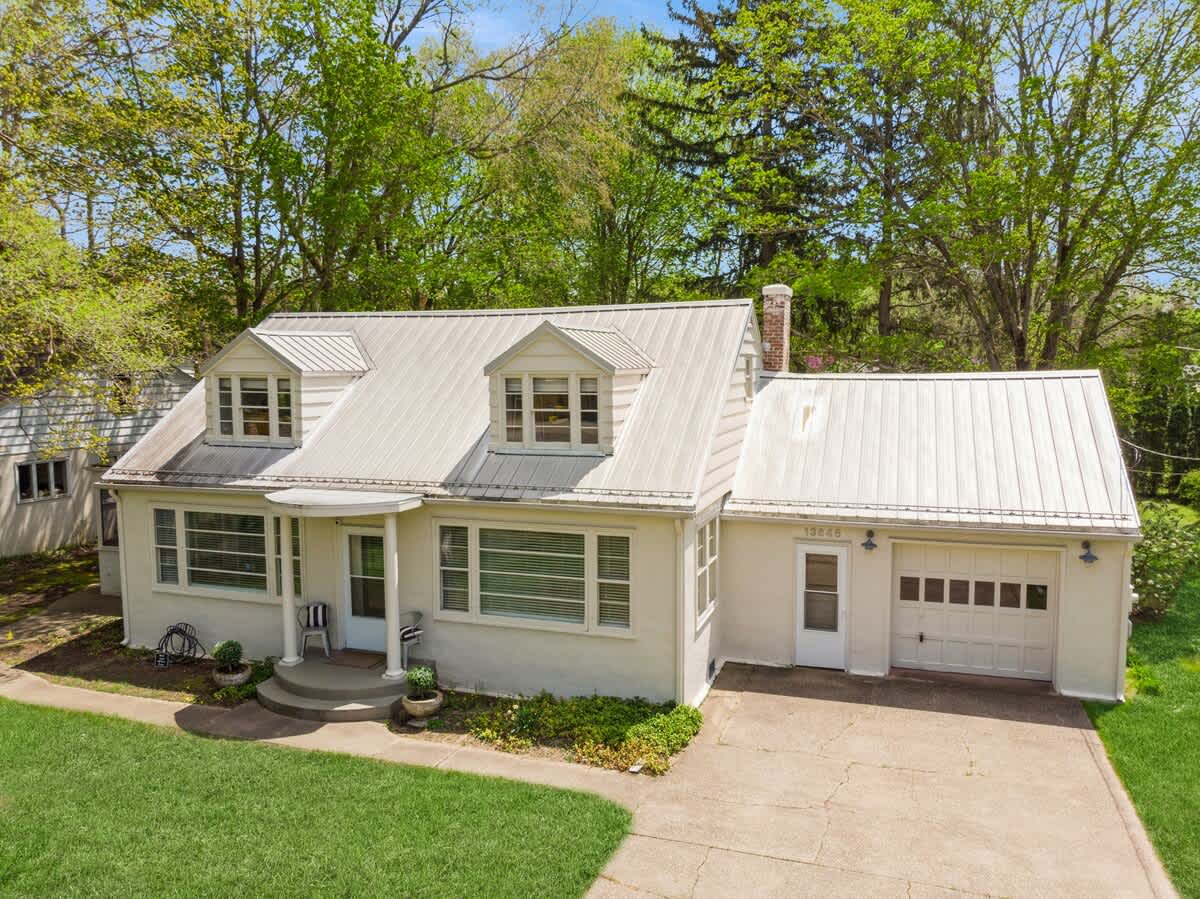 Angled front view of the white cottage, highlighting the attached garage with a white panel door, a concrete driveway, and the light-colored metal roof with dormer windows.