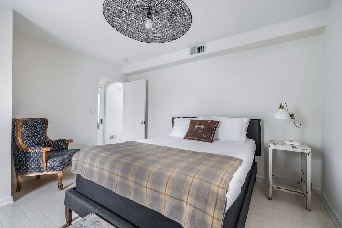 A bright bedroom with white wooden floors and walls, featuring a dark gray upholstered bed with white bedding and a plaid throw, a patterned armchair, and a unique black woven ceiling light fixture.
