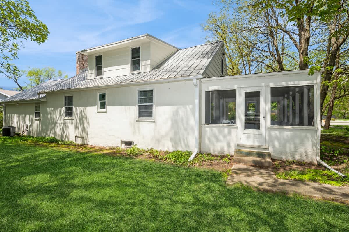 Side view of the white house with a textured stucco-like finish, featuring a screened porch addition with a white door and steps, and a well-maintained green lawn under a bright sky.