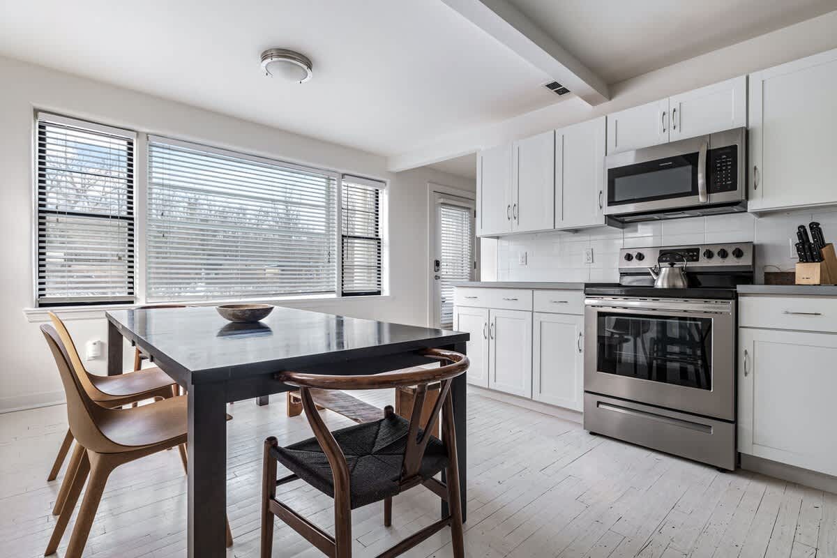 Integrated kitchen and dining space featuring white cabinets, stainless steel appliances, a dark wood dining table with various chairs, and large windows providing natural light.