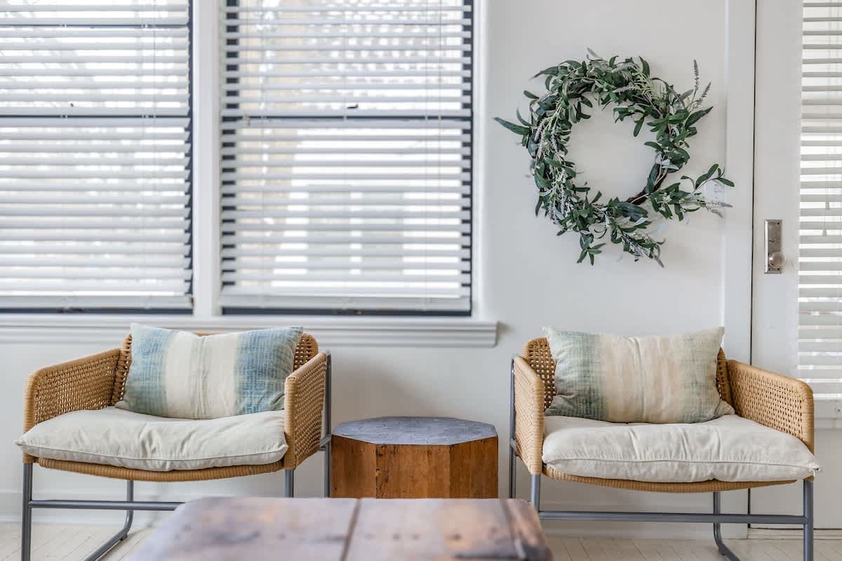 Close-up of two woven armchairs with light cushions and striped pillows, flanking a small hexagonal wooden side table. A decorative wreath hangs on the white wall next to a window with blinds.